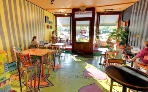Colorful café interior with striped walls, vibrant tables, and chairs, featuring a welcoming entrance and a few patrons.