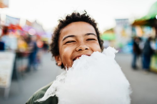 A joyful child smiles widely while holding a large cotton candy at a fair, with colorful stalls in the background.