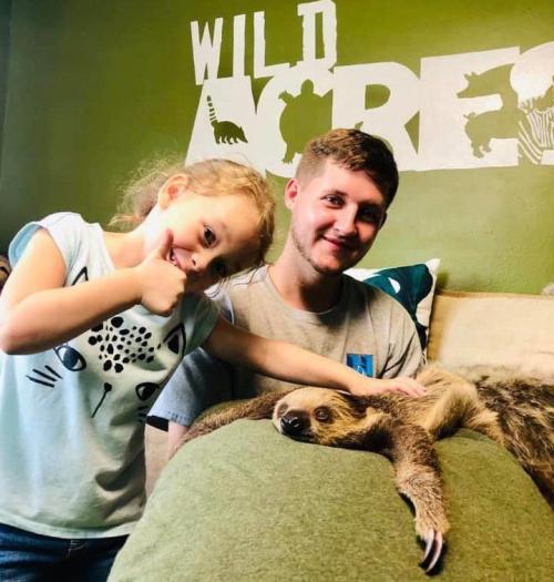 A young girl gives a thumbs up next to a smiling young man, both sitting with a sloth on a couch.