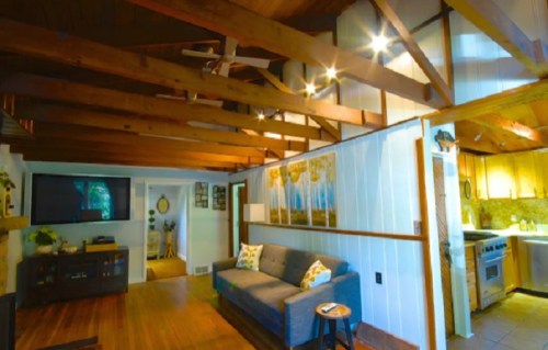 Cozy living room with wooden beams, a gray sofa, and a kitchen visible in the background. Warm, inviting atmosphere.