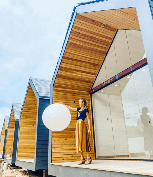 A woman in a blue top and yellow skirt holds a white lantern outside modern wooden houses with sloped roofs.