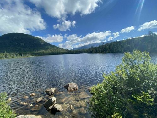 A serene lake surrounded by lush greenery and mountains under a partly cloudy sky. Rocks line the water's edge.
