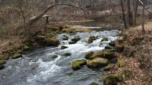 A serene stream flows over moss-covered rocks, surrounded by trees and a calm pond in a wooded area.