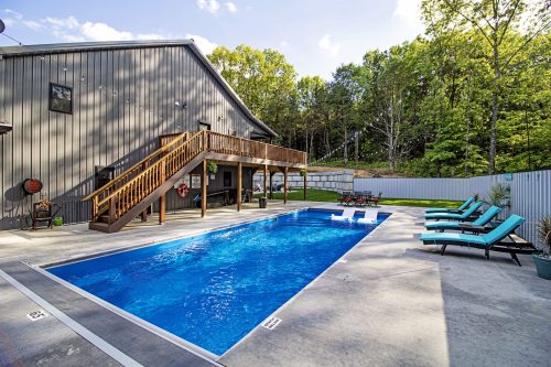 A clear blue pool surrounded by lounge chairs, with a wooden deck and a building in a wooded area.