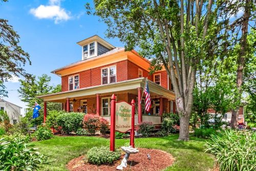 A charming red brick inn surrounded by greenery, featuring a welcoming sign and flags in a picturesque setting.