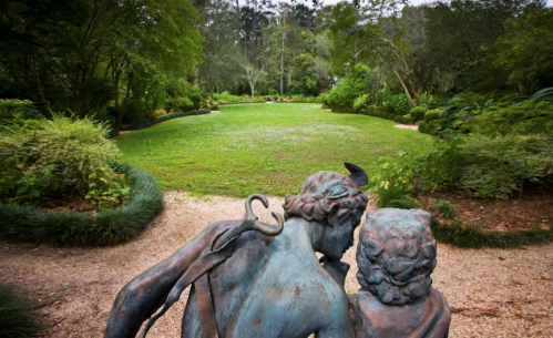 A bronze statue of two figures in a lush garden, overlooking a green lawn surrounded by trees and plants.