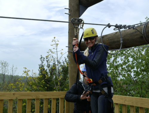 A woman in a helmet prepares to zip line, while a person assists her from the platform. Trees are visible in the background.