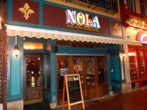 Exterior of "NOLA on the Square" restaurant, featuring colorful signage and a welcoming entrance with a chalkboard menu.