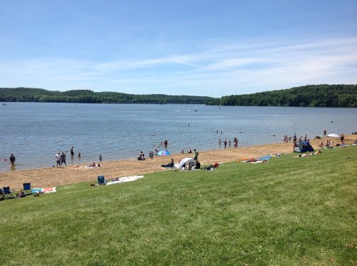 A sunny beach scene with people swimming and relaxing on the sand, surrounded by green hills and a calm lake.