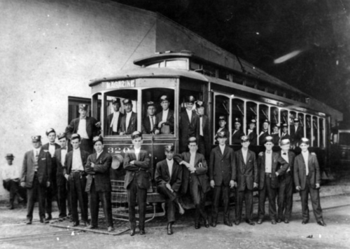 A historic black-and-white photo of a streetcar with a large group of men, some in uniforms, posing in front of it.