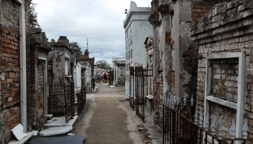 A narrow path between weathered tombs in a cemetery, with brick walls and wrought iron fences, under a cloudy sky.