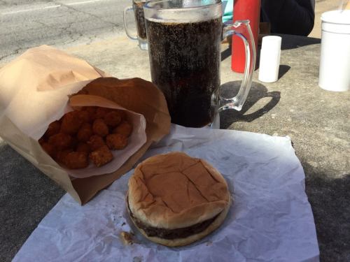 A burger on a paper wrapper, a bag of tater tots, and a large glass of soda on a table outdoors.