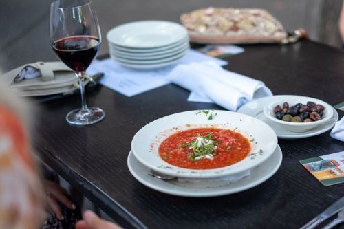 A bowl of red soup garnished with herbs, accompanied by a glass of red wine and a plate of olives on a table.