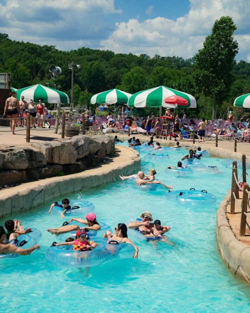 A busy water park with people floating on tubes in a lazy river, surrounded by green and white umbrellas.