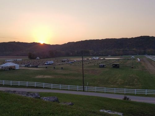 Sunset over a grassy field with tents and a white fence, surrounded by hills and a clear sky.