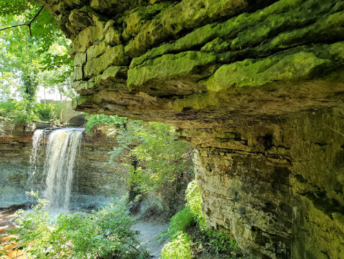 A lush green landscape featuring a waterfall cascading over rocky cliffs, surrounded by trees and foliage.