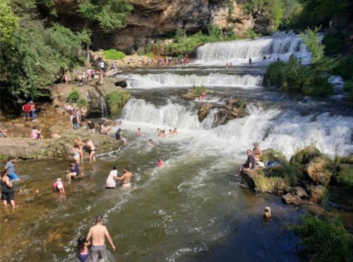 People enjoying a sunny day by a waterfall, playing in the water and relaxing on the rocks. Lush greenery surrounds the scene.