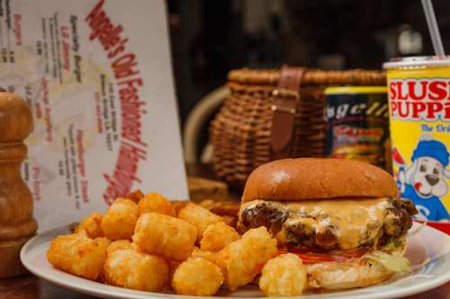 A plate featuring a cheeseburger with lettuce and tomato, served with crispy tater tots and a drink in the background.