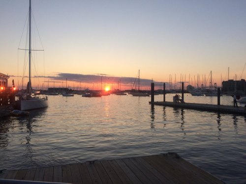 Sunset over a marina with sailboats and reflections on the water, creating a serene evening atmosphere.