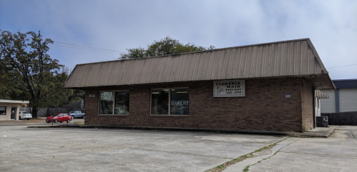 A brick building with a metal roof, featuring a bakery sign and large windows, set in a parking lot.
