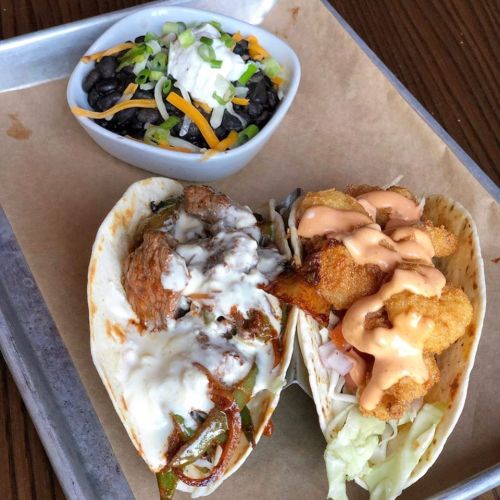 Two tacos filled with meat and shrimp, topped with sauce, served with a side of black beans and cheese.