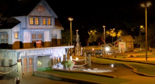 A brightly lit mini-golf course at night featuring a rustic mill and surrounding greenery.