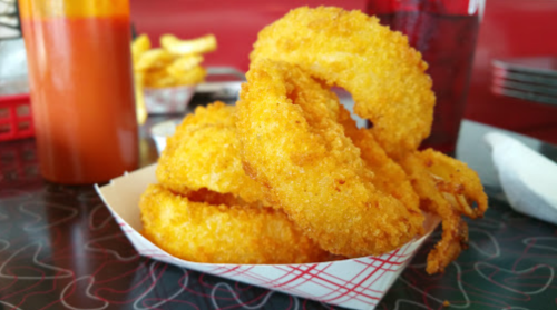A basket of golden, crispy onion rings with a side of dipping sauce, set on a patterned table.