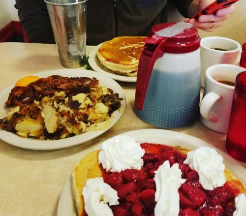 A table with breakfast items: pancakes, bacon, hash browns, and a pitcher of syrup, alongside coffee and orange slices.