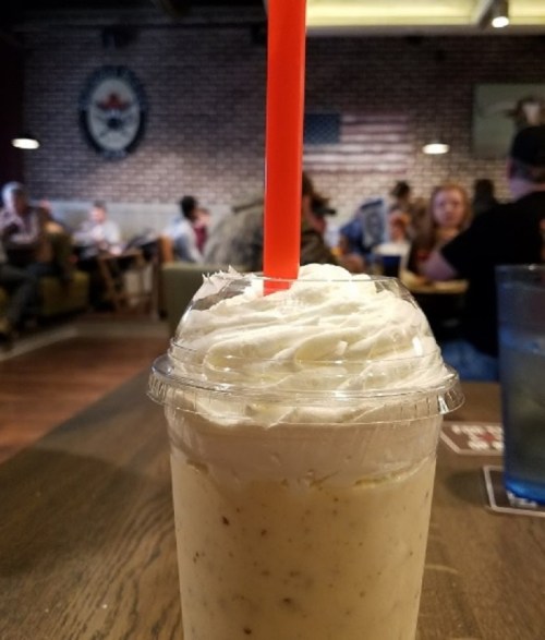 A close-up of a milkshake with whipped cream and a red straw, set on a wooden table in a busy restaurant.