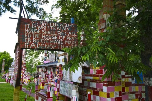 Colorful, handmade sign with biblical text, surrounded by vibrant, decorated structures and lush greenery.
