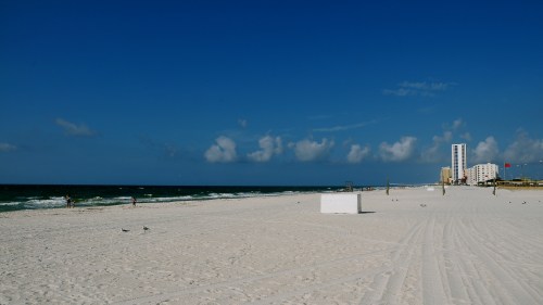 A sunny beach scene with white sand, gentle waves, and a distant building under a clear blue sky.