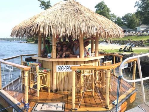 A group of people enjoying drinks at a tiki bar by the water, surrounded by trees and a sunny atmosphere.