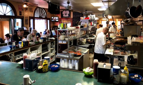A busy diner kitchen with chefs cooking, customers dining, and a counter filled with condiments and utensils.