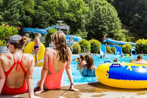 Two people in swimsuits sit by a pool, while others enjoy water slides and floaties in a sunny, green setting.