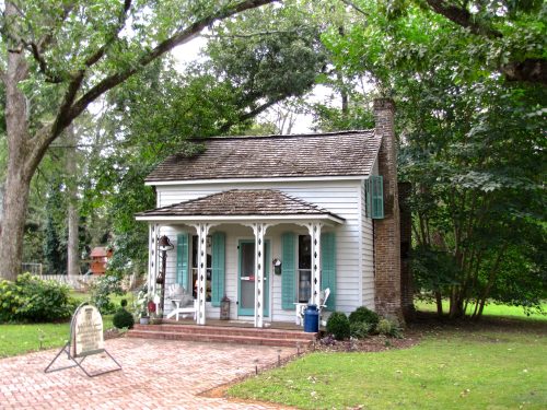 A charming white cottage with a shingled roof, green shutters, and a brick pathway, surrounded by lush greenery.