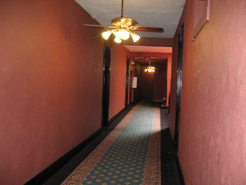 A long hallway with red walls, a patterned carpet, and ceiling fans, leading to a doorway at the end.