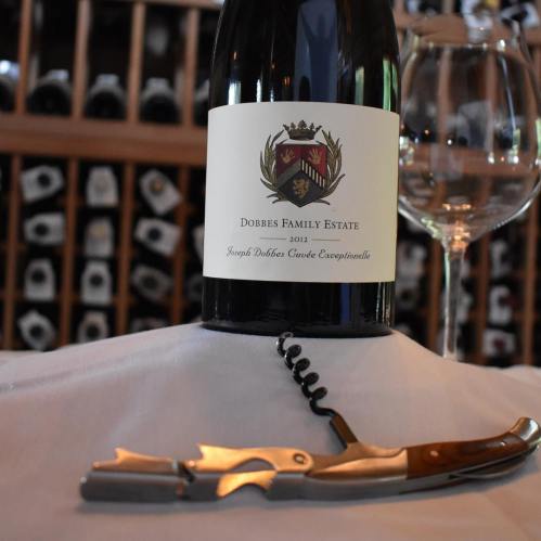 A bottle of Dobbes Family Estate wine with a corkscrew and wine glass on a table, set against a wine rack backdrop.
