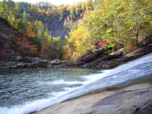 A serene river flows over rocks, surrounded by vibrant autumn foliage and steep cliffs in a tranquil natural setting.