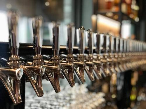 A row of shiny beer taps lined up on a bar, with blurred bottles in the background.