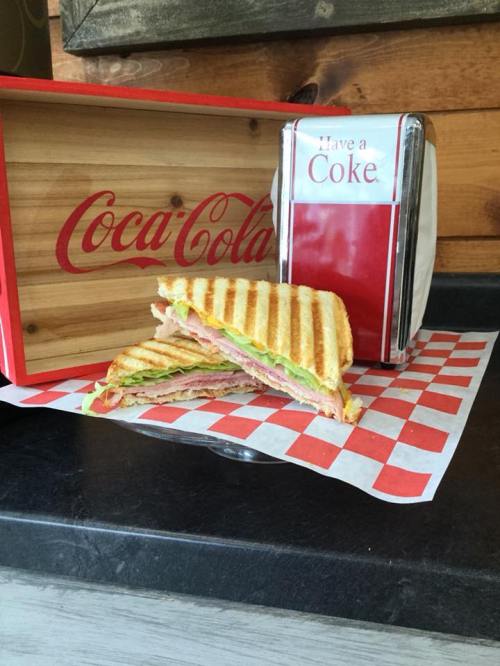 A toasted sandwich with layers of ham, lettuce, and tomato, served with a Coca-Cola box and a retro drink dispenser.