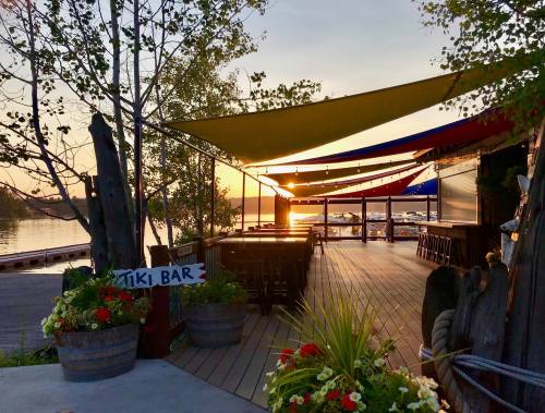 A tiki bar by the water at sunset, featuring colorful awnings and flower pots, with boats in the background.