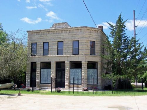 Historic two-story stone building with large windows, surrounded by trees and a fence, under a clear blue sky.