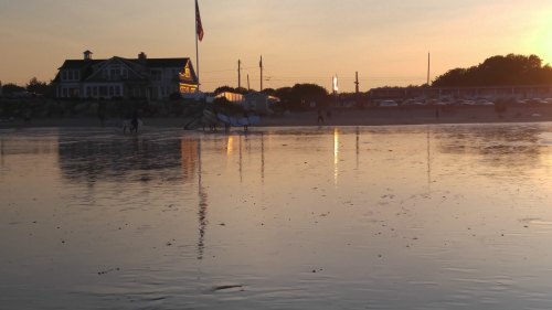 A beach at sunset with a house, American flag, and people walking along the water's edge. Reflections in the wet sand.