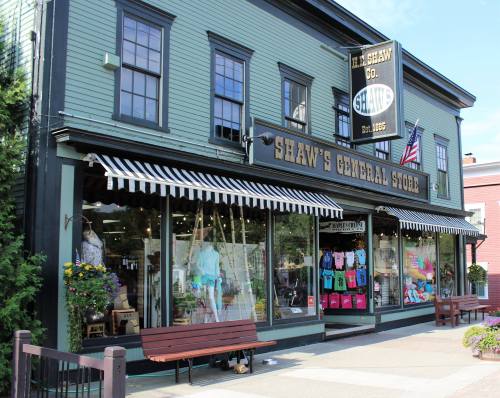 A charming green general store with striped awnings, displaying colorful clothing and flowers outside.