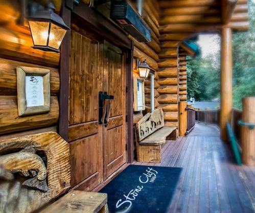 Entrance to a rustic log cabin with wooden doors, benches, and a welcome mat, surrounded by nature.