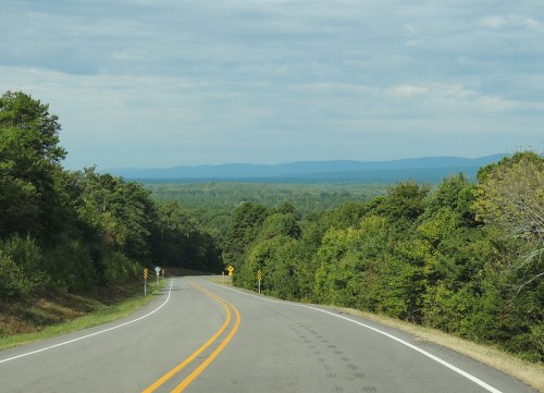 A winding road through lush green trees, leading to distant blue mountains under a cloudy sky.
