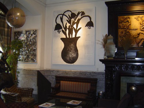 A large black and white painting of a flower vase hangs on a wall in a stylish, decorated room with elegant furniture.