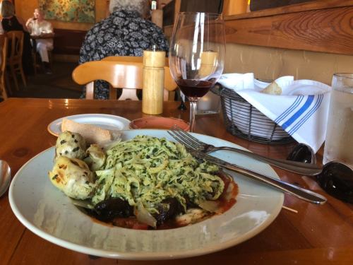A plate of pasta with greens, artichokes, and sun-dried tomatoes, accompanied by a glass of red wine and a bread roll.