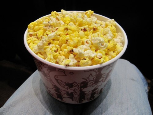 A large cup of buttery popcorn resting on a person's lap, with a patterned design on the cup.