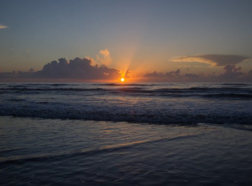 A serene beach scene at sunset, with the sun dipping below the horizon and clouds reflecting warm colors in the sky.